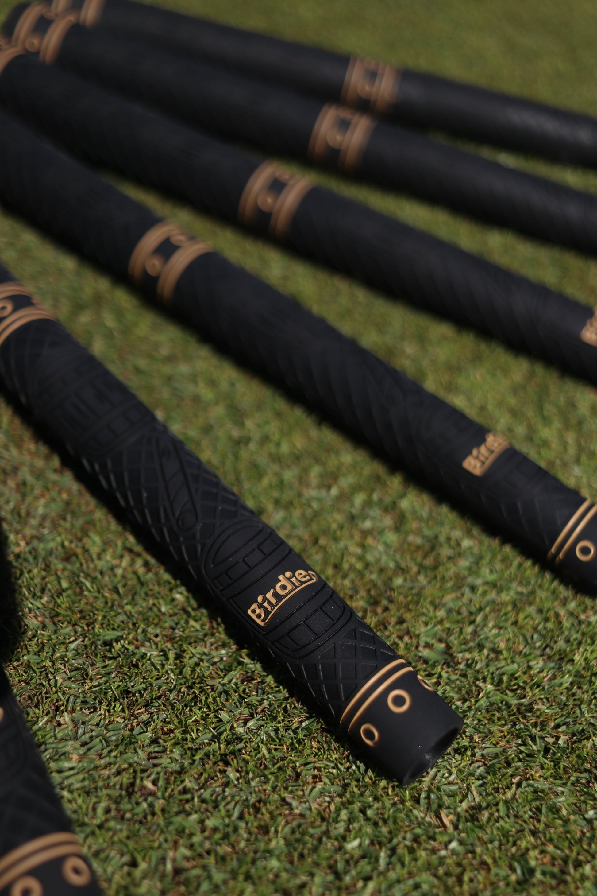 Birdie Golf Grips Smoke & Barrel black golf club grips with raised texture and gold detailing on a green grass field.